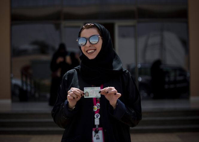 Eine Frau mit locker sitzendem Kopftuch, Sonnenbrille und lackierten Fingernägeln zeigt stolz ihren neu erworbenen Führerschein.