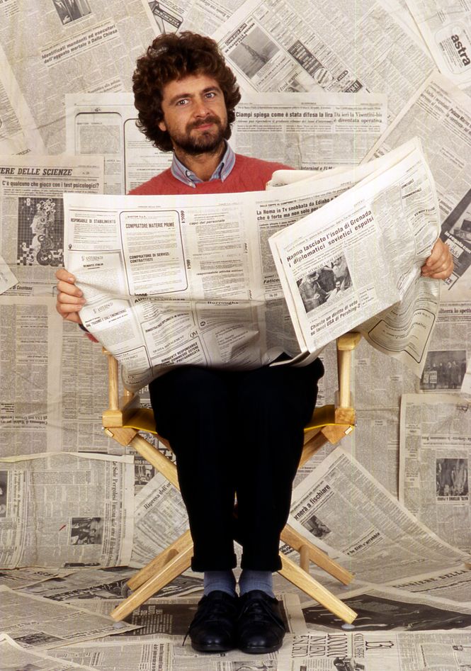 Grillo sitzend und Zeitung lesend vor einer Wand, die ganz mit Zeitungen zugeklebt ist