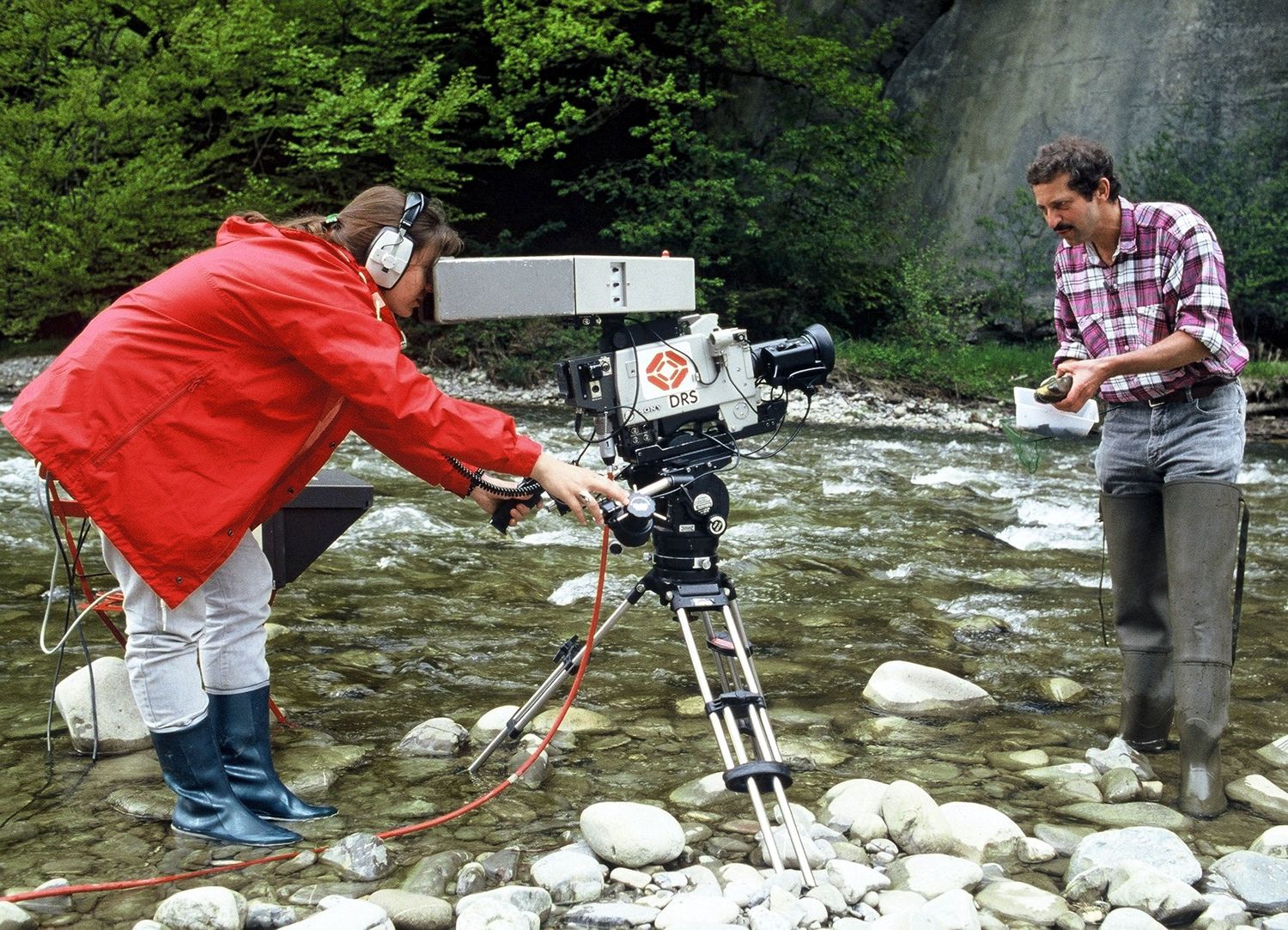 Kamerafrau steht im Wasser eines Flusses und filmt einen Fischer