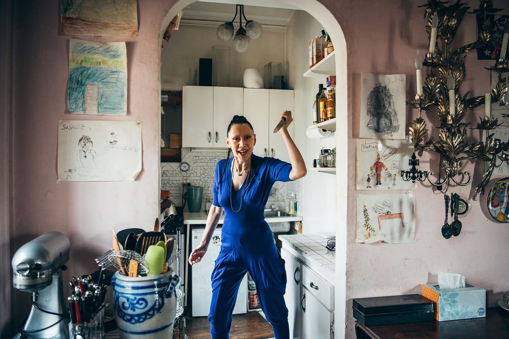 Susanne Bartsch posiert in einem blauen Overall mit gezücktem Küchenmesser in ihrer Küche.