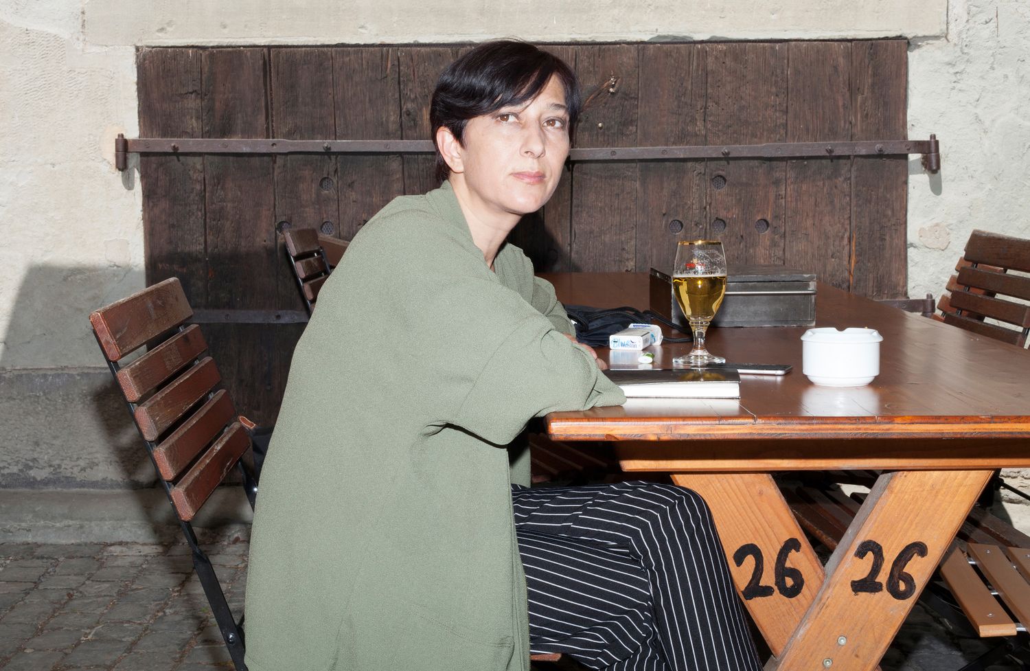 Portrait von Esen Isik, sie sitzt an einem Tisch vor einem Bier, Zigaretten und Notizbuch