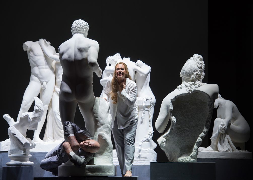 Szene in der Glyphothek mit antiken Skulpturen. Alviano in glänzendem Anzug und verstümmelten Händen zwischen den Füssen einer männlichen Skulptur eingeklemmt. Carlotta Nardi in weissem Hemd und weisser Hose. 
