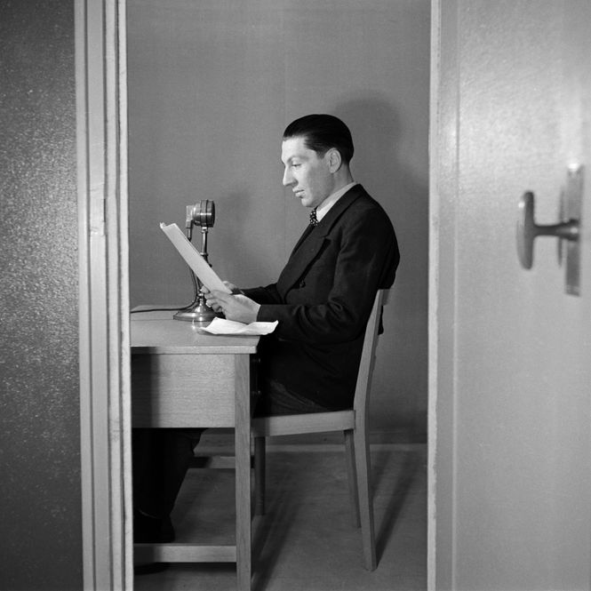 Ein Journalist sitzt in einem kleinen Raum vor einem Mikrofon und liest von einem Blatt