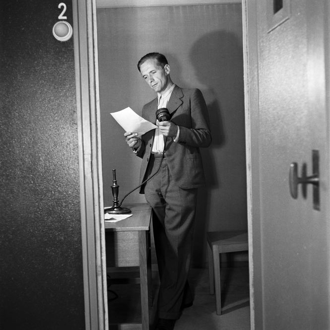 Ein Jounalist steht in einem kleinen Raum mit einem Mikrofon und liest von einem Blatt
