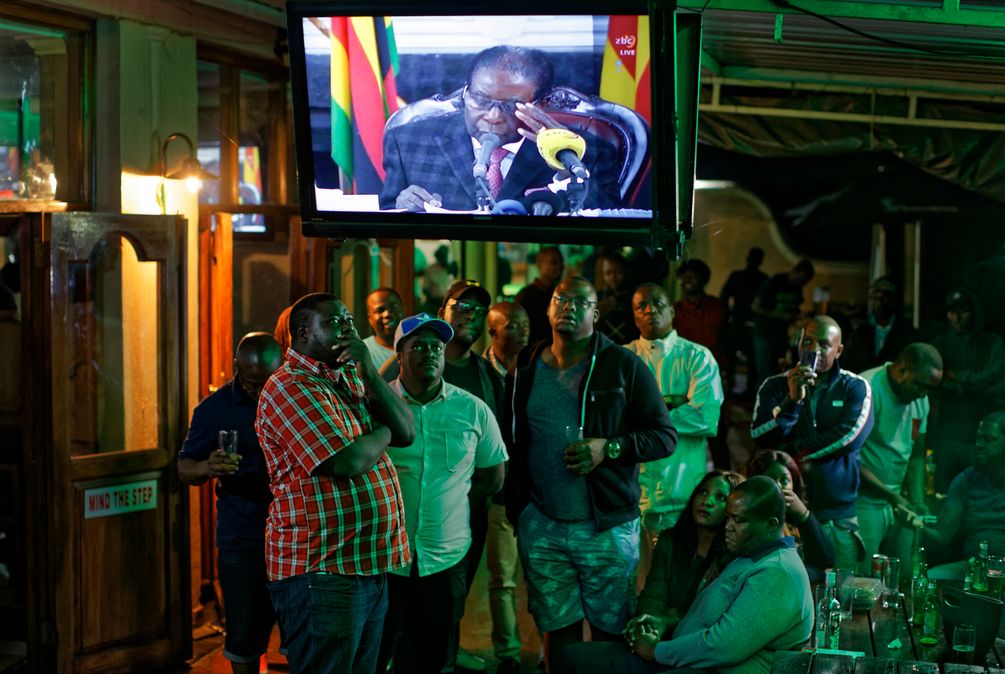 eine Gruppe von Menschen schaut auf einen TV, wo Mugabe an einem Rednerpult sitzt