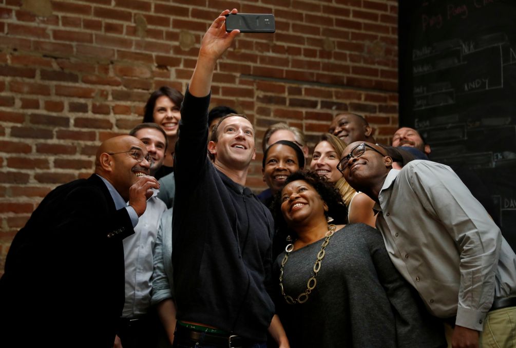 Mark Zuckerberg steht inmitten einer Gruppe von lächelnden Menschen und macht mit ihnen ein Selfie